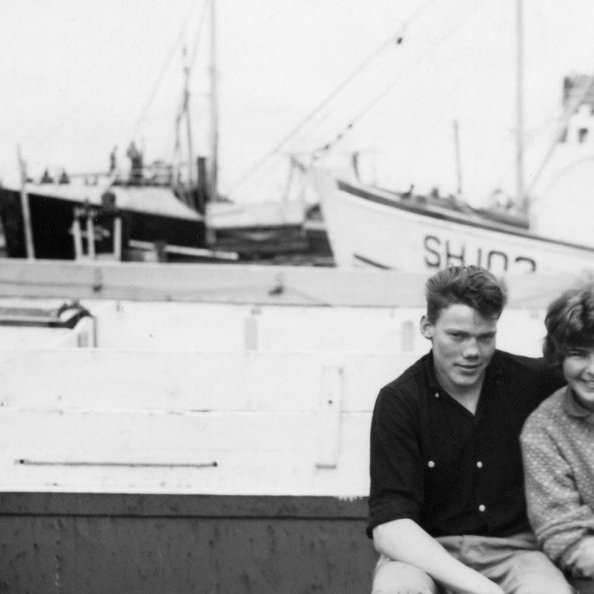 Þessi skemmtilega mynd er sennilega tekin á Siglufirði. Ungi maðurinn á myndinni er Ívar Árnason en okkur vantar nafnið á konunni. Líklega er þetta Sigurfari frá Grundarfirði þar sem skráningarnúmerið sést að hluta til og eflaust þekkja einhverjir hvort það er rétt og kunna einnig skil á hinum bátunum.