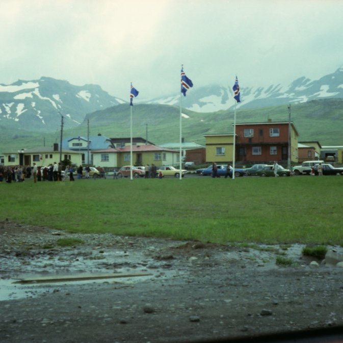 Á þessari mynd líklega 17 júní eftir rigningunni að dæma og íslenski fáninn er dreginn að húni. Túnið þarna á mótum Grundargötu 30 og Hrannarstígs 7 var kallaður skrúðgarður þótt aldrei hafi verið á þessum árum sett niður tré eða blóm, en þarna fór iðulega fram ýmis hátíðarhöld eins og á 17 júní og Þrettándanum. Þessi mynd er tekin tekin eftir 1980 og á myndinni má sjá að ýmis uppbygging er í gangi lengst til vinstri má sjá húsnæði Búnaðarbankans í byggingu og fremst á myndinni liggur spýta og holræsisrör sem krakkar sem voru að alast upp á þessum tíma kunnu að nota í ýmsa leiki. Í dag er kominn trjágróður og blóm á milli Grundargötu 30 og heilsugæslunnar.