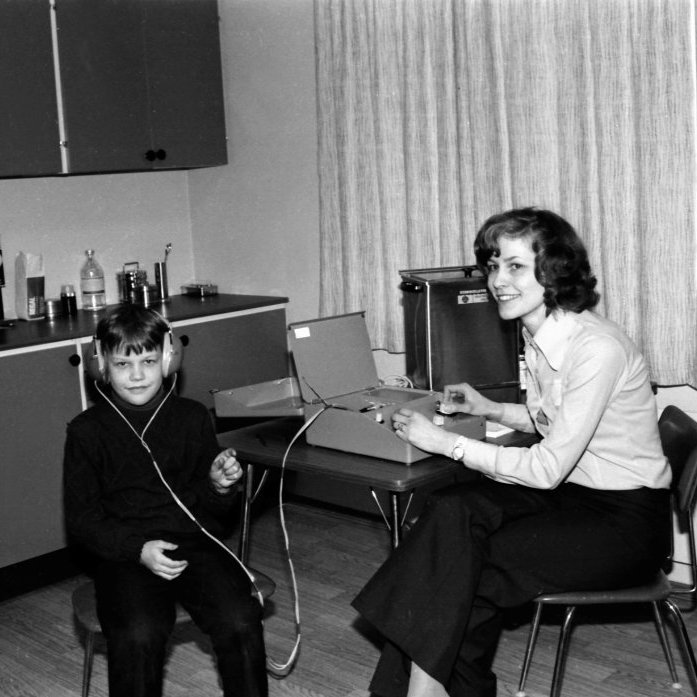 Hildur Sæmundsdóttir ljósmóðir að störfum, í heilbrigðisstéttinni unnu bæði karlmenn og kvennmenn á þessum árum þótt læknarnir hafi í flestum tilfellum þá verið karlmenn. Hildur er að gera heyrnarmælingu á Sveinbirni Halldórssyni með nýju tæki heilsugæslunnar.