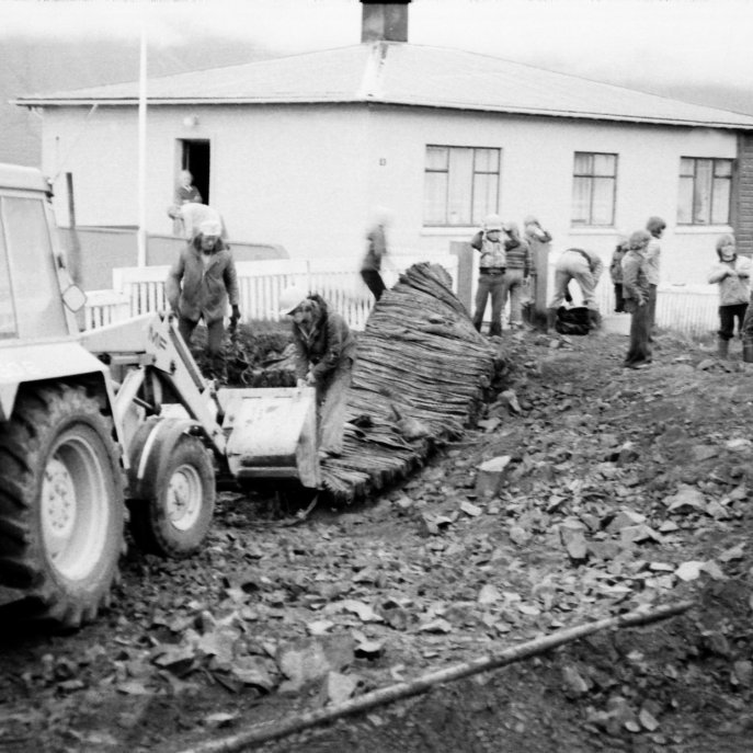 Vinna við gatnagerð á Grundargötunni, hér er verið að sprengja fyrir framan Grundargötu 8 og fylgist húsmóðirn Halla Halldórsdóttir ásamt Hrefnu Hjartardóttur með mönnunum vinna og með krökkunum. Gæti verið Elis Guðjónsson á gröfunni? Rafn Ólafsson og ungur maður sem vantar nafn á eru að vinna við sprengju mottuna. Þarna eru nokkrir krakkar að fylgjast með og læra hvernig á að byggja upp gatnakerfi, það væri gaman að fá nöfn þeirra.